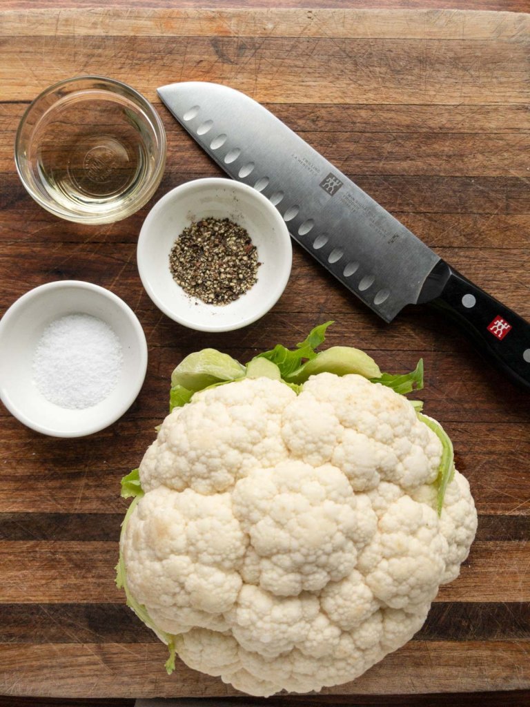 Head of cauliflower, chefs knife, oil in a glass bowl, salt in a white bowl, pepper in a white bowl.  Ingredients sitting on a brown cutting board. 