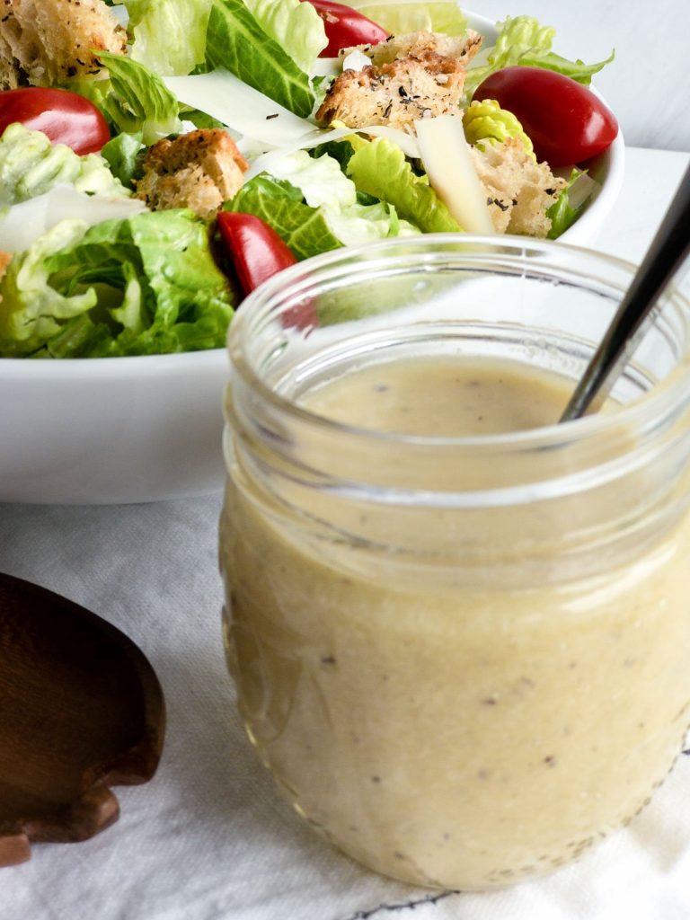 Caesar salad dressing in a glass jar next to white bowl that contains romaine lettuce, cherry tomatoes, croutons and grated Parmesan cheese
